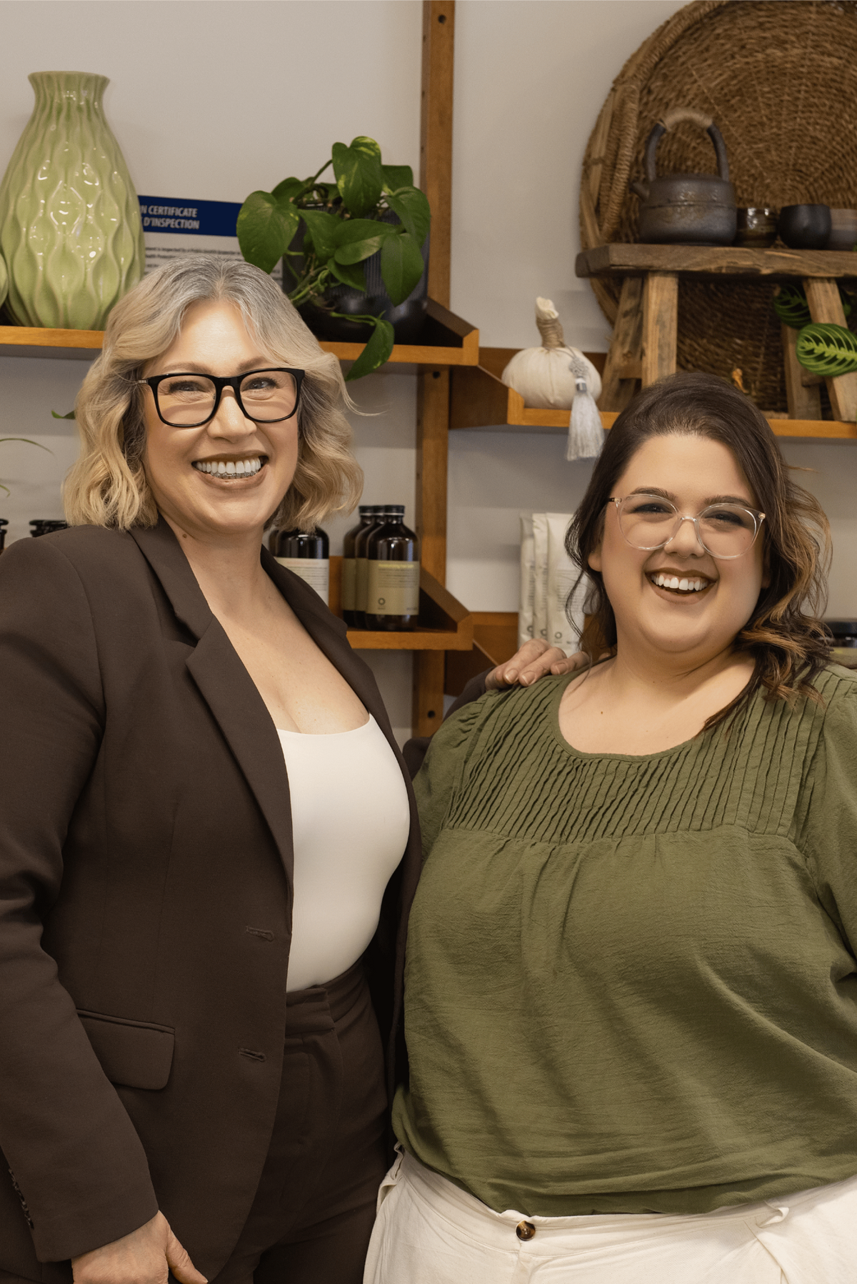 Two women smiling and posing together in a cozy, styled indoor setting with wooden shelves, plants, and home decor. The woman on the left wears a dark brown blazer and pants over a white top, while the woman on the right wears an olive green short-sleeve top and cream wide-leg pants.